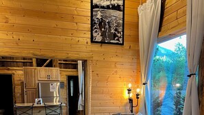 L’intérieur chaleureux d’un chalet avec des murs en bois, un coin repas comprenant une table et des chaises, un canapé gris agrémenté d’un coussin décoratif et une fenêtre avec des rideaux.