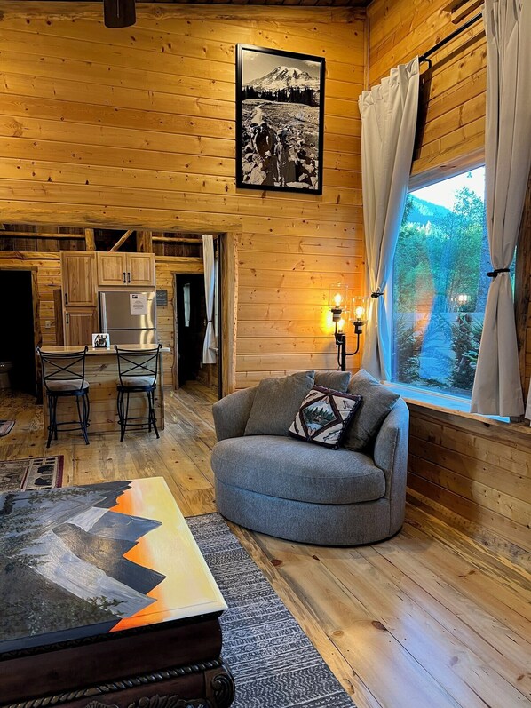 A cozy cabin interior with wooden walls, a dining area with a table and chairs, a gray sofa with a decorative pillow, and a window with curtains.