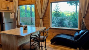 L’intérieur d’une cabane est aménagé avec du bois, il comprend un coin cuisine avec un réfrigérateur, une table à manger avec des chaises et une grande fenêtre avec des rideaux.
