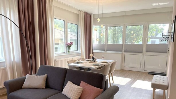 Una sala moderna con un sofá gris, una mesa de comedor con sillas y ventanas grandes con cortinas.