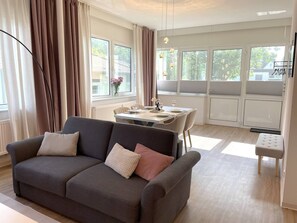 Ein modernes Wohnzimmer mit einem grauen Sofa, einem Esstisch mit Stühlen und großen Fenstern mit Vorhängen.