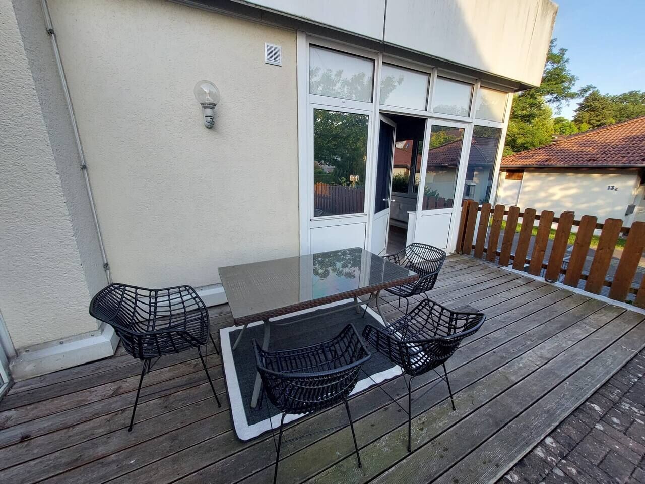 Un patio con un tavolo in vetro e quattro sedie nere, un pavimento in legno e un edificio bianco con una finestra e un'entrata.