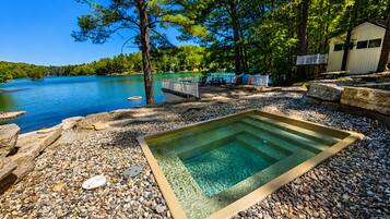 A swimming pool with a pebble surround, a wooden deck, and a lake view.