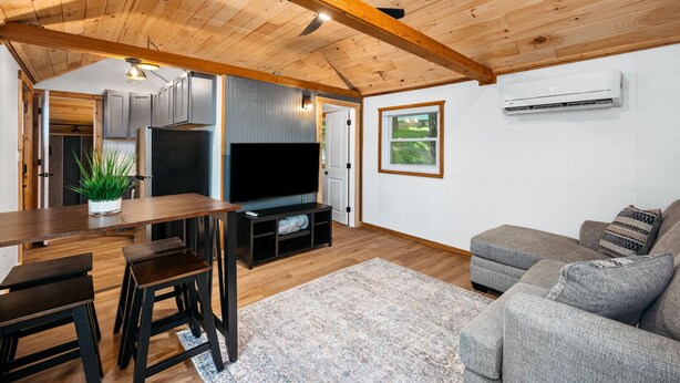 A modern living room with a wooden ceiling, a flat-screen TV, a grey sofa, and a dining area with a wooden table and chairs.