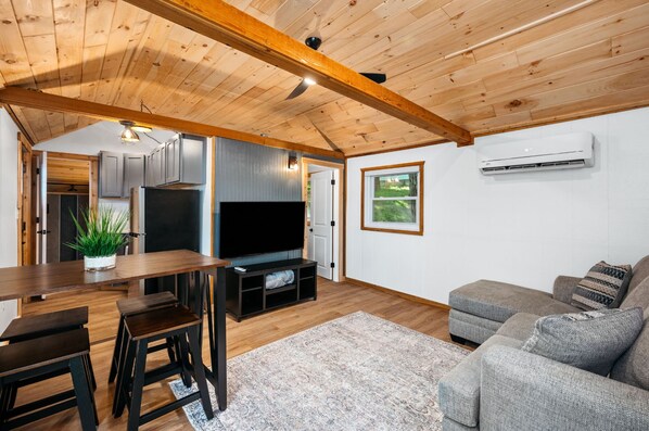Una sala de estar moderna con techo de madera, un televisor de pantalla plana, un sofá gris y un comedor con mesa y sillas de madera.