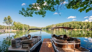 Two pontoon boats docked at a pier with a calm lake and forested shoreline in the background.