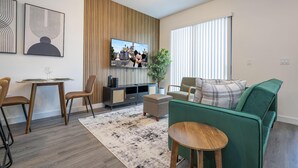 A modern living room with a green sofa, wooden coffee table, and a TV on a wooden panel wall. - 3232 - Beautiful 3br Home near Disney - The Hub at Westside (Kissimmee)