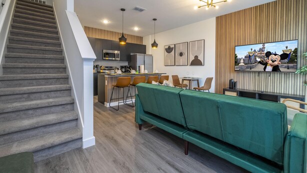 A modern kitchen with a central island, a dining area with chairs, a green sofa, and a staircase. - 3232 - Beautiful 3br Home near Disney - The Hub at Westside (Kissimmee)