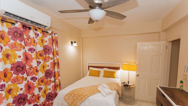 A hotel room with a bed, a ceiling fan, an air conditioner, a nightstand with a lamp, and a wooden dresser.