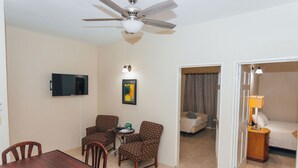 A hotel room with a dining table, two patterned armchairs, a TV, a ceiling fan, a bedside lamp, and a bed with a nightstand.