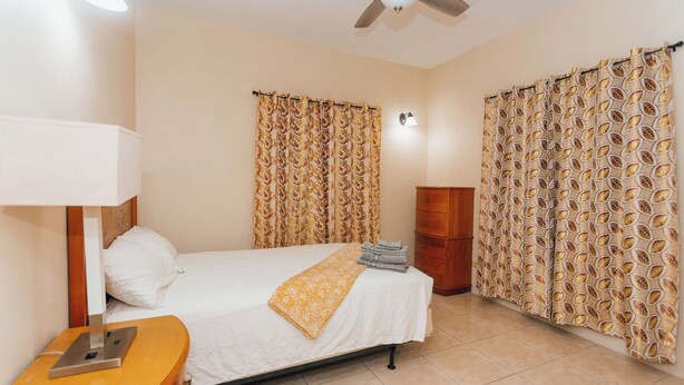 A hotel room with a bed, a nightstand, a lamp, a ceiling fan, and curtains with a leaf pattern.