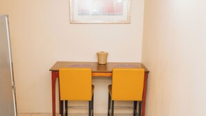 Un pequeño comedor con una mesa de madera y dos sillas amarillas, un cubo de basura color beige y un cuadro en la pared.