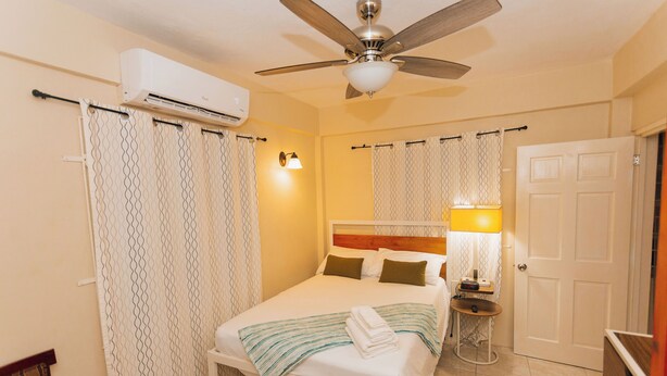 Un dormitorio con cama, ventilador de techo, equipo de aire acondicionado, mesita de noche con lámpara y una puerta.