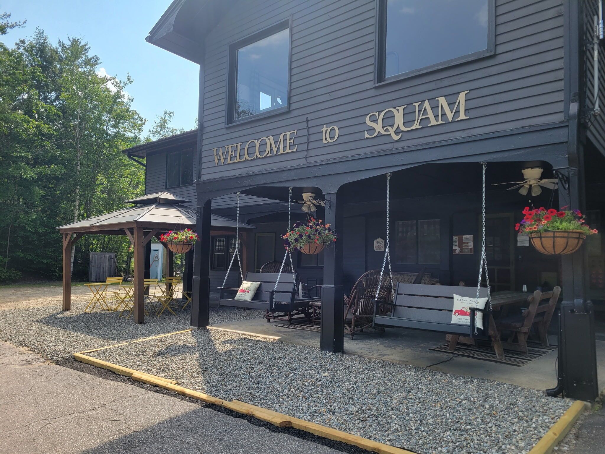 Un bâtiment sur lequel on peut lire « BIENVENUE À SQUAM », avec des sièges extérieurs dotés de balançoires et des jardinières suspendues remplies de fleurs.