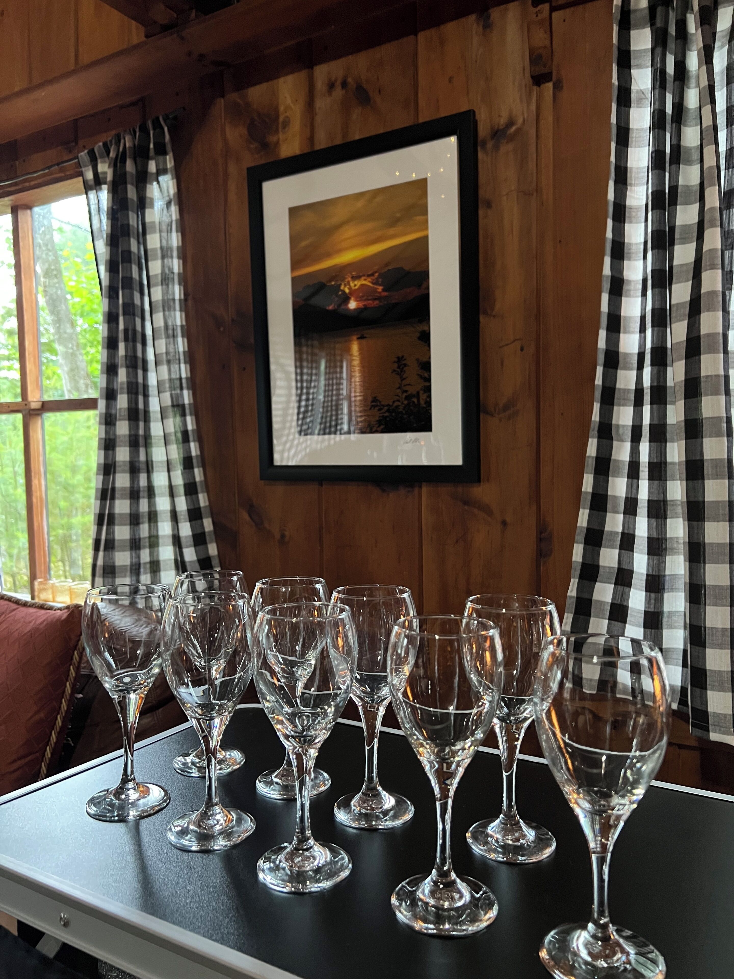 A table with multiple wine glasses arranged in a row, a framed picture on the wall, and checkered curtains on the windows.