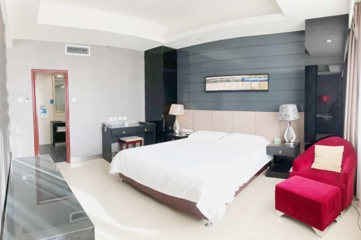A modern hotel room with a large bed, a desk, a red armchair, and a small table.
