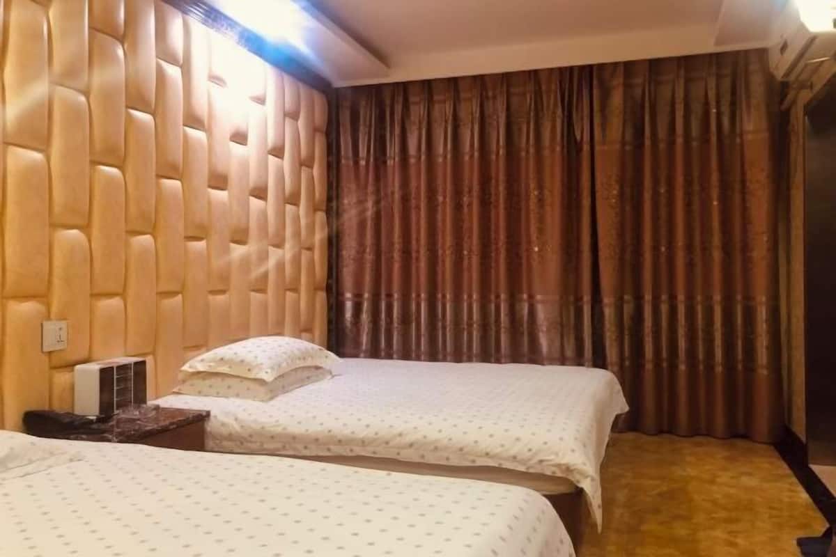 Habitación de hotel con un cabecero capitonado, dos camas con ropa blanca, alfombra marrón y un amplio ventanal con cortinas.