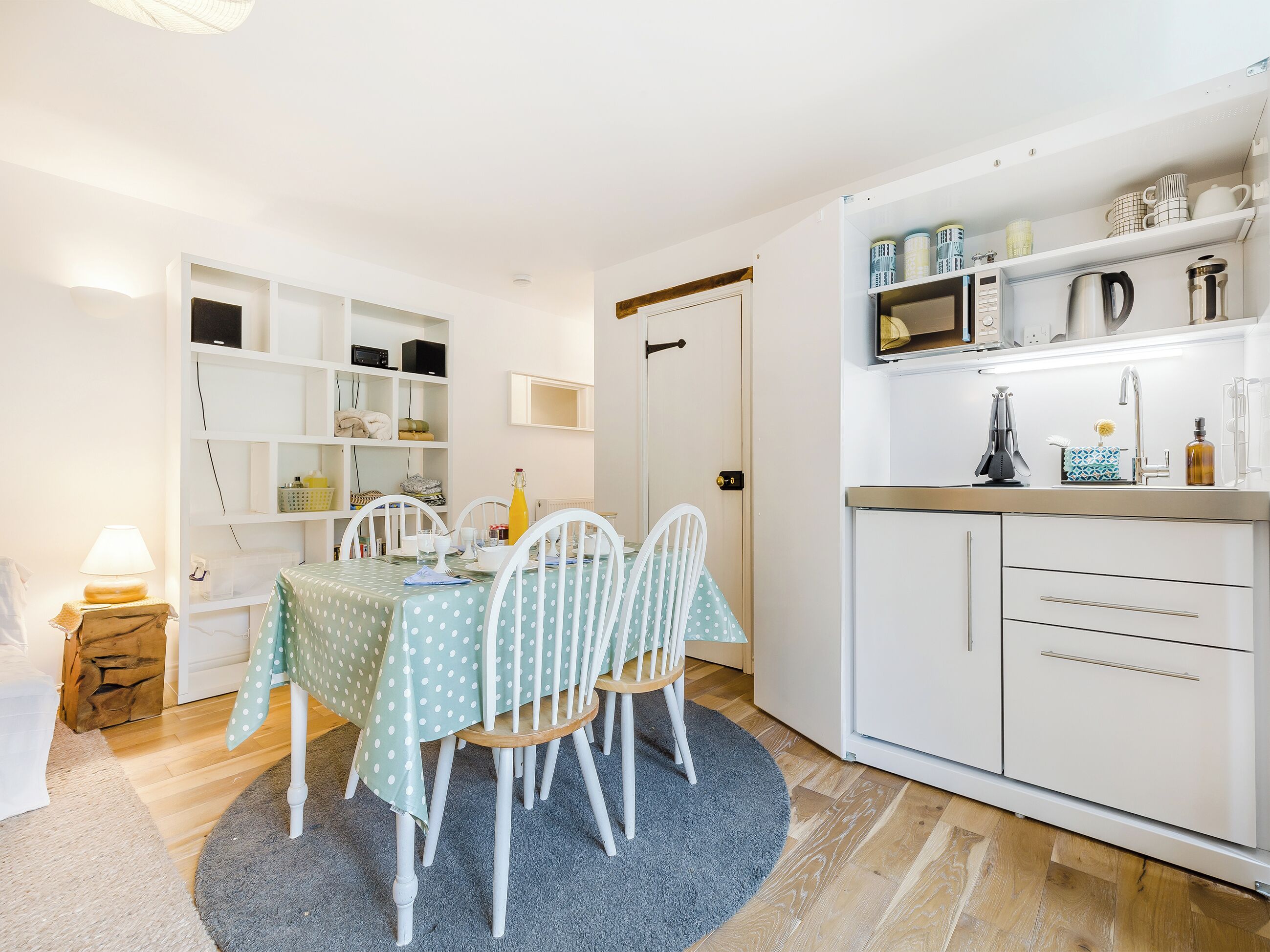 A compact kitchen with white cabinets, a dining table with chairs, and a small side table with a lamp.