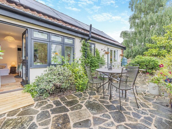 Een terras van stenen met een tafel en stoelen, een wit hek en een tuin met verschillende planten.