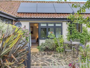 Een huis met een stenen terras, een tuin met diverse planten, een zonnepaneel op het dak en een raam met uitzicht op de tuin.