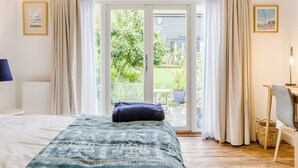 Um quarto com cama, janela com cortinas, radiador, luminária e vista para um jardim.