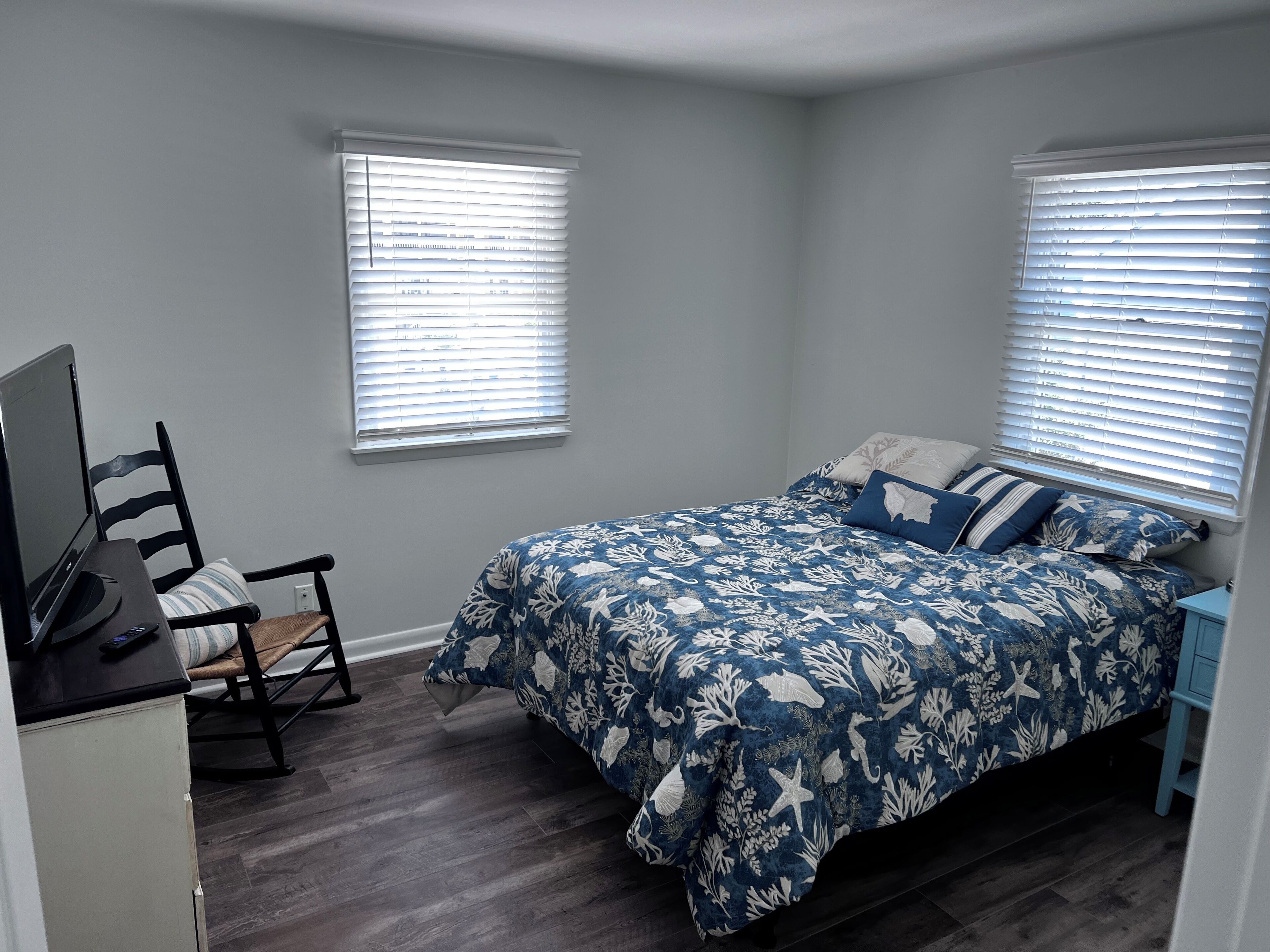 A bedroom with a bed, two windows with blinds, a desk with a chair, and a dresser.