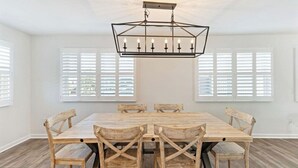 A dining room with a rectangular wooden table and six chairs, a geometric chandelier, and large windows with white shutters.