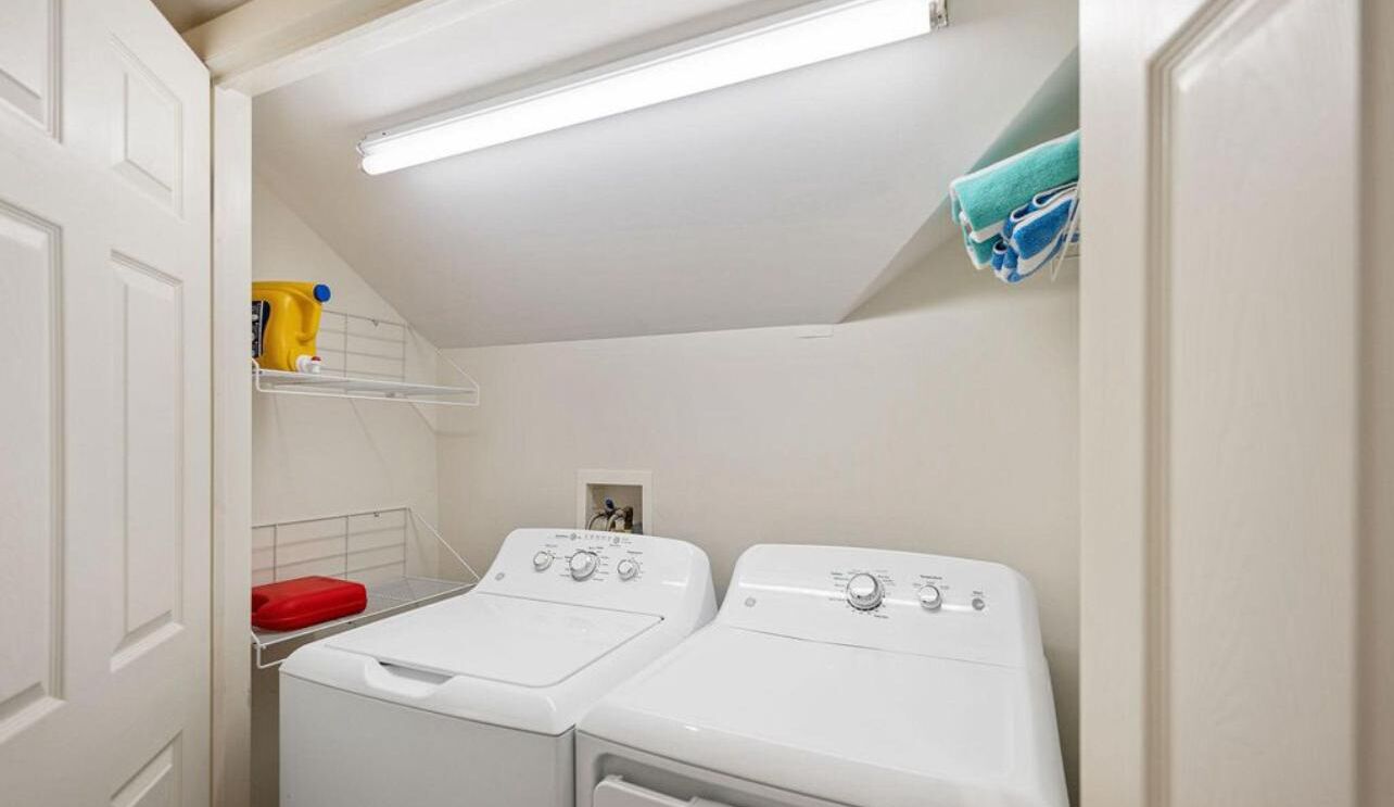 A laundry room with two white front-loading washing machines, a red laundry basket, and a shelf with cleaning supplies.