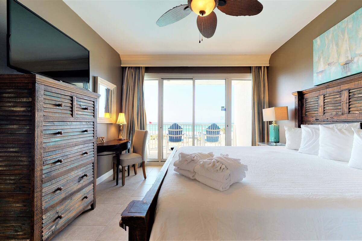 A hotel room with a large bed, a wooden dresser, a desk with a lamp, and a view of the ocean through a sliding door.