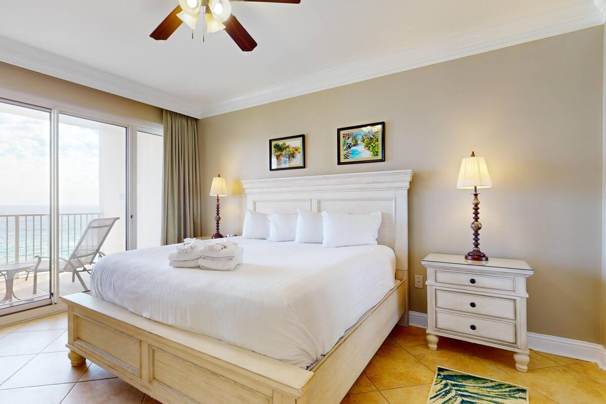 A hotel room with a large bed, two bedside lamps, a nightstand, a chair, and a view of the ocean.