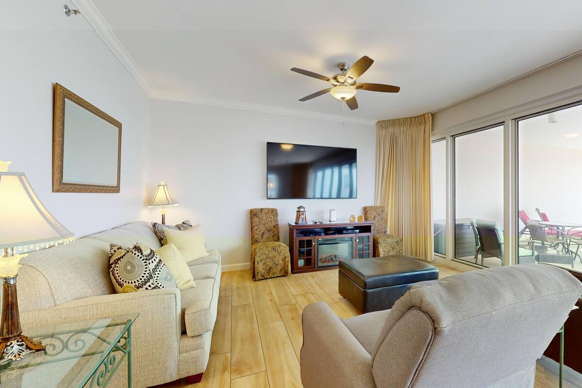 A spacious living room with a ceiling fan, a flat-screen TV, and a large window offering a view of the outdoors.