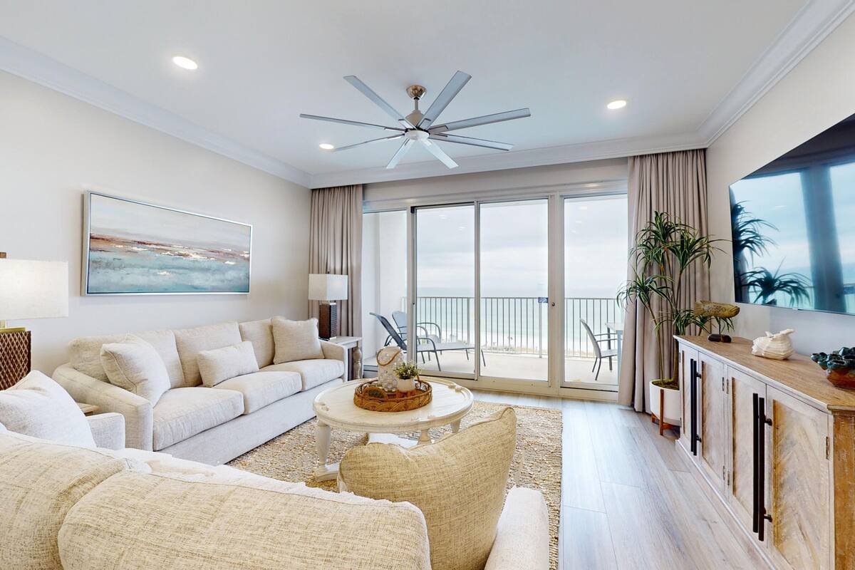 A spacious living room with a large sofa, a coffee table, and a TV. There's a balcony with a view of the ocean.