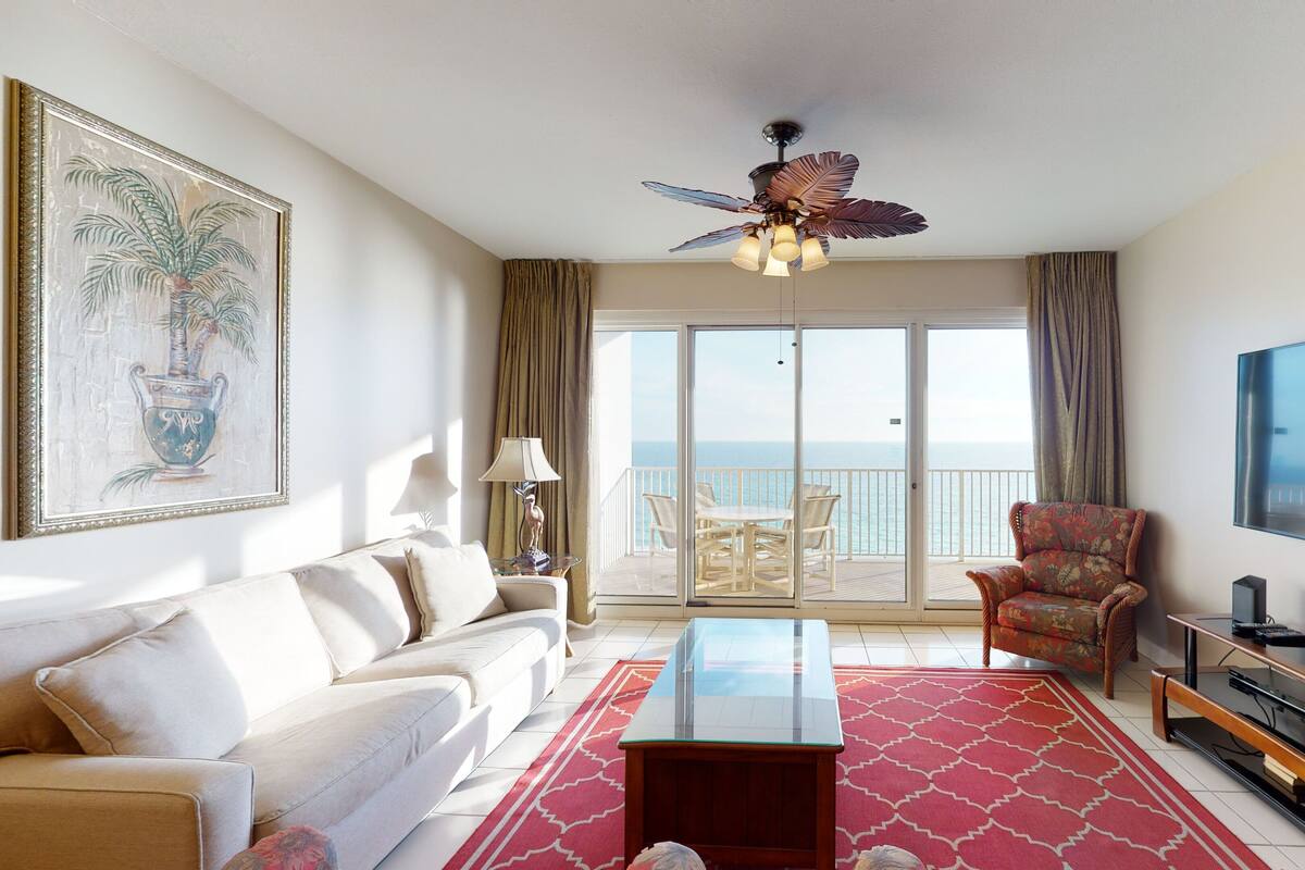 A living room with a sofa, a glass coffee table, a TV, a chair, and a balcony with a view of the sea.