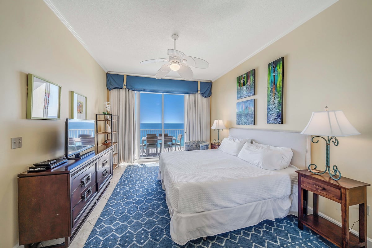 A hotel room with a bed, a desk with a lamp, a TV, a balcony with a view, and artwork on the walls.