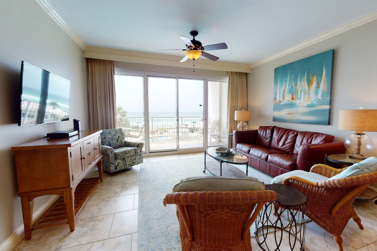 A spacious living room with a balcony, a leather sofa, wicker chairs, a TV, and a painting.