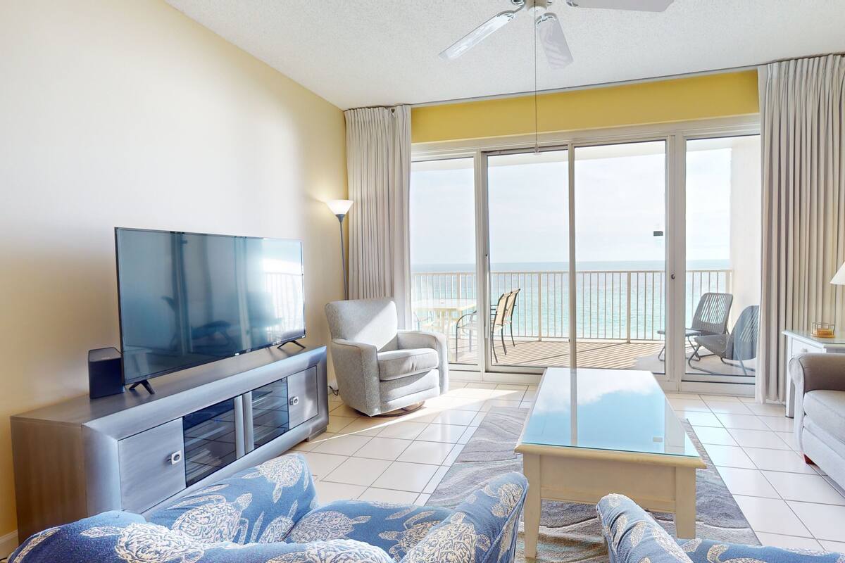 A modern living room with a flat-screen TV, a sofa, a coffee table, and a balcony with a view of the sea.