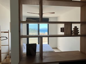 Een kamer met een houten plankensysteem, een bed met een beddegoed met patroon, en uitzicht op het strand en de bergen door een groot raam.