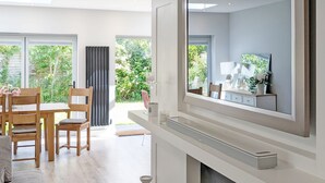 A modern living room with a large mirror, a dining area with wooden furniture, and a view of a garden through the windows. - A family house that sleeps 6 guests  in 3 bedrooms (West Wittering)
