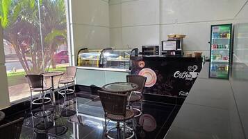 A modern coffee shop with a black counter, wicker chairs, and a large window offering a view of palm trees and a street.