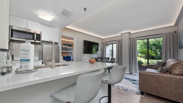 A modern kitchen with white cabinets, a stainless steel microwave, and a large island with bar stools. - Luxury 2 Suites, Near Parks and Int'l Drive (Orlando)
