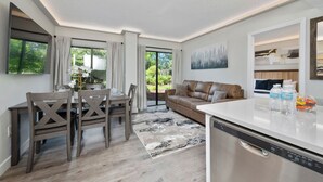 A modern kitchen with a dishwasher, a dining area with a table and chairs, a sofa, and a large window with curtains. - Luxury 2 Suites, Near Parks and Int'l Drive (Orlando)