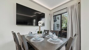 A dining area with a wooden table set for four, white plates, bowls, and a vase with white flowers. There is a large flat-screen TV mounted on the wall above the table. - Luxury 2 Suites, Near Parks and Int'l Drive (Orlando)