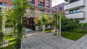 En udendørs patio med et pergolaoverdækning, grønt og udsigt til moderne bygninger i baggrunden.