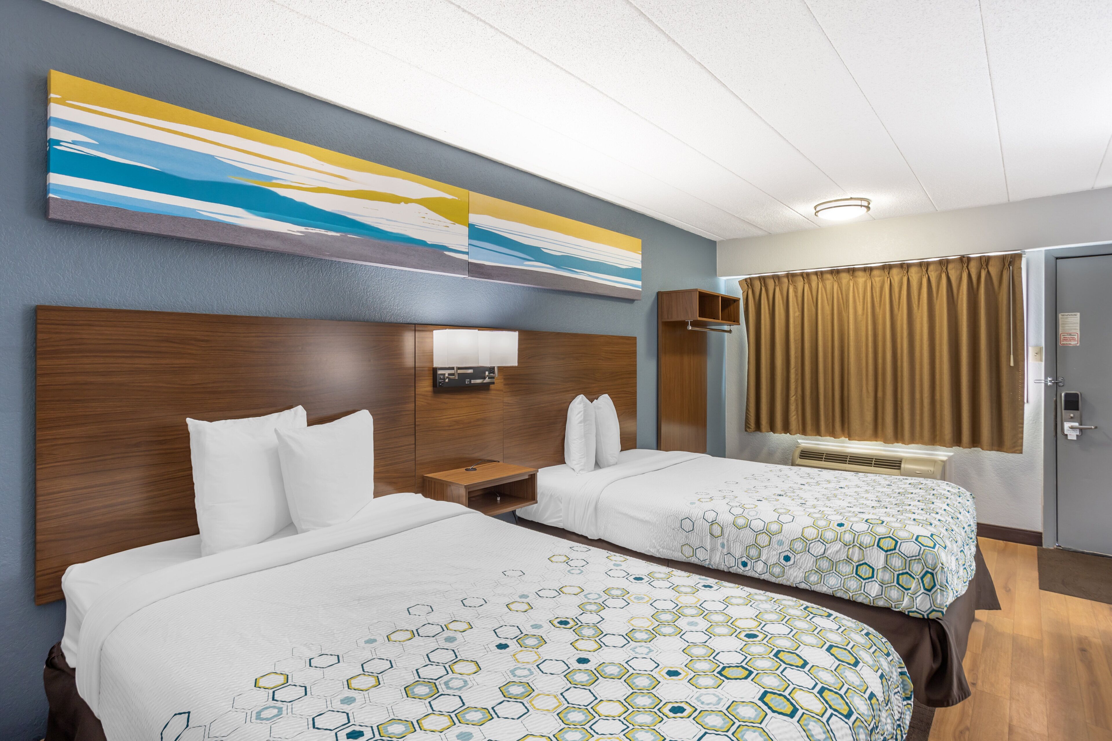 A hotel room with two beds, a nightstand, a wall-mounted lamp, a window with curtains, and a door.