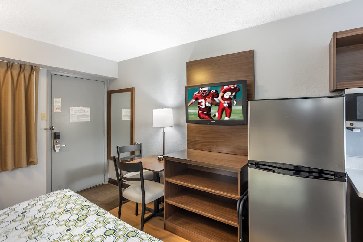 Habitación de hotel con cama, escritorio con silla, refrigerador y televisor de pared mostrando un partido deportivo.