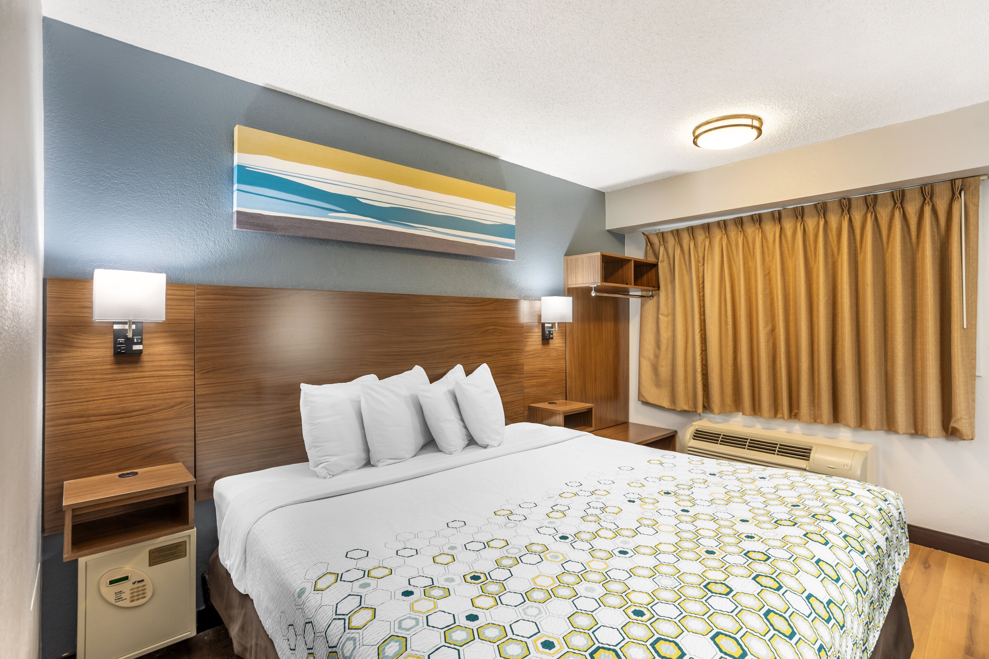 A hotel room with a bed, a nightstand, a wall-mounted lamp, a window with curtains, and a ceiling light.