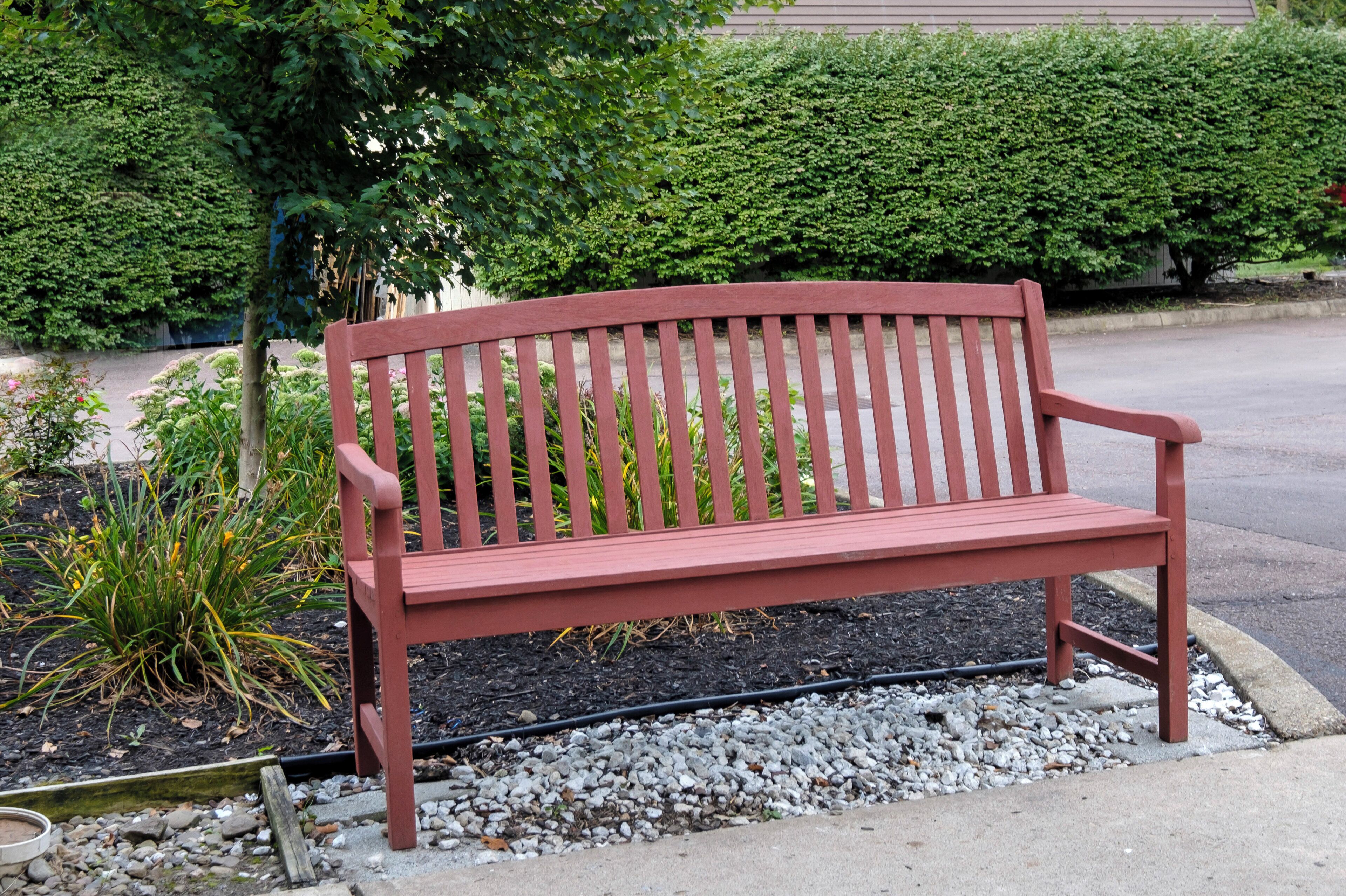 A red bench with a backrest and armrests, positioned on a gravel path with a hedge and plants in the background.