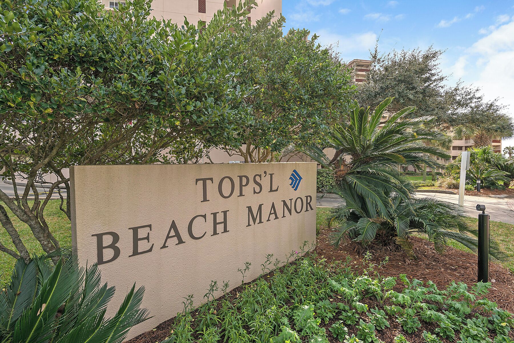 A sign for Topsl Beach Manor with a view of surrounding greenery and buildings.