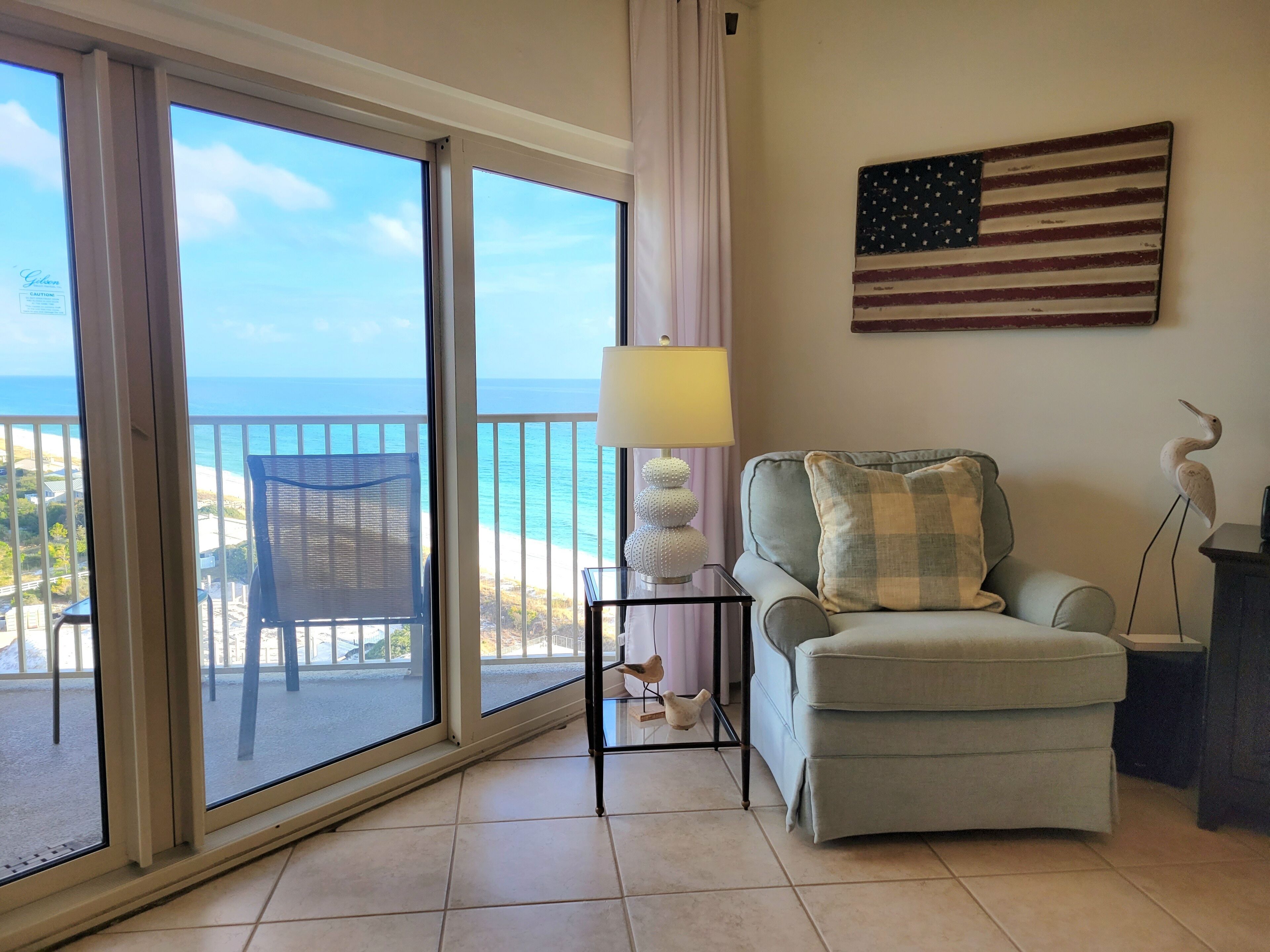Una habitación con vistas a la playa, una silla, una lámpara, una mesita y una estatua decorativa de un pájaro.