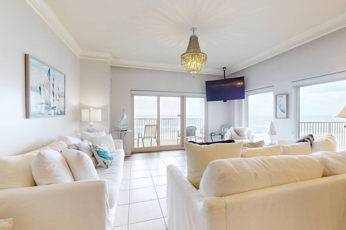 A spacious living room with white furniture, a chandelier, and a view to the outside.